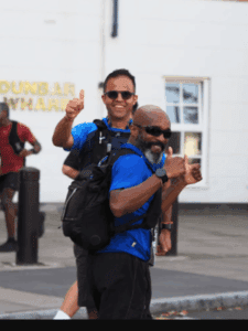 two men wearing matching blue shirts and black shorts both with thumbs up happy expressions running